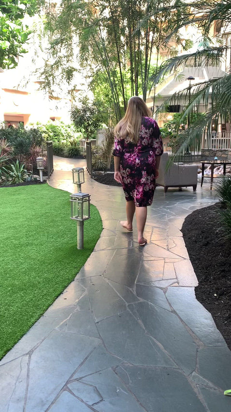 blonde size 18 woman wearing a straight dress with draped top and billow sleeves starts out with her back to camera and then walks towards camera on a path in a garden