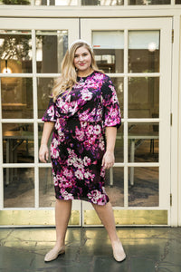 Tall blonde size 18 woman wears a straight dress with a draped top, and cross pleated top and billow sleeves. The print is purple and white flowers with a black background  